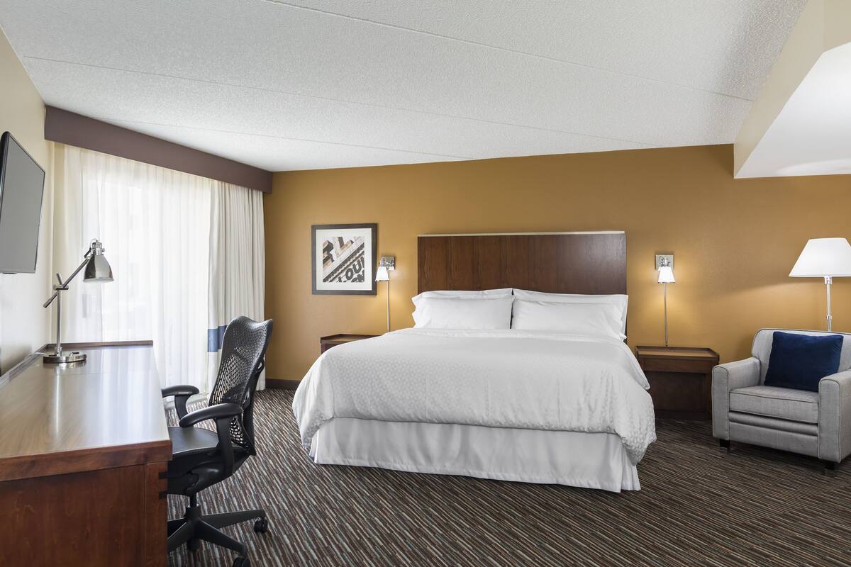 A hotel room with a large bed, a desk with a chair, a small armchair, and a TV on the wall.