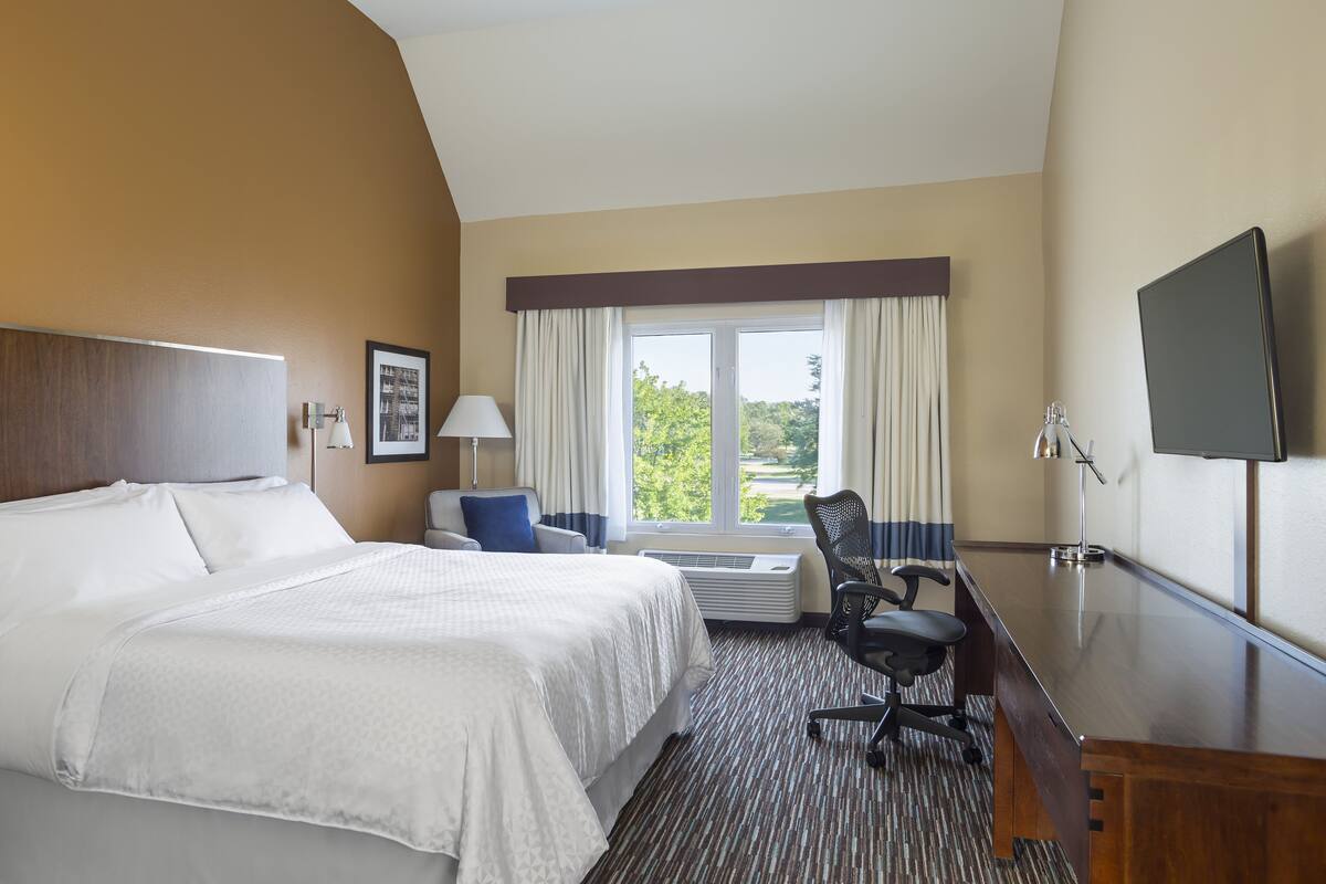 A hotel room with a large bed, a desk, a chair, a TV, and a window with curtains.