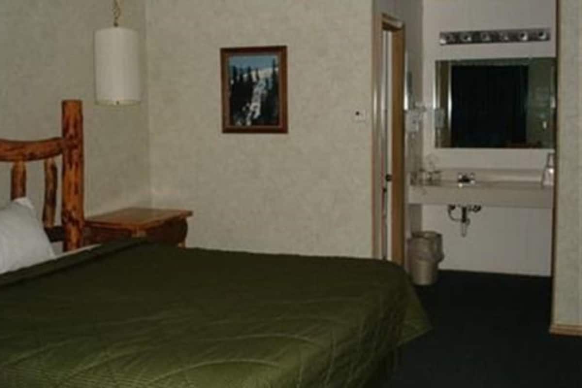 Quarto de hotel com cama, mesa de cabeceira de madeira, quadro na parede, banheiro com pia e espelho, e janela.