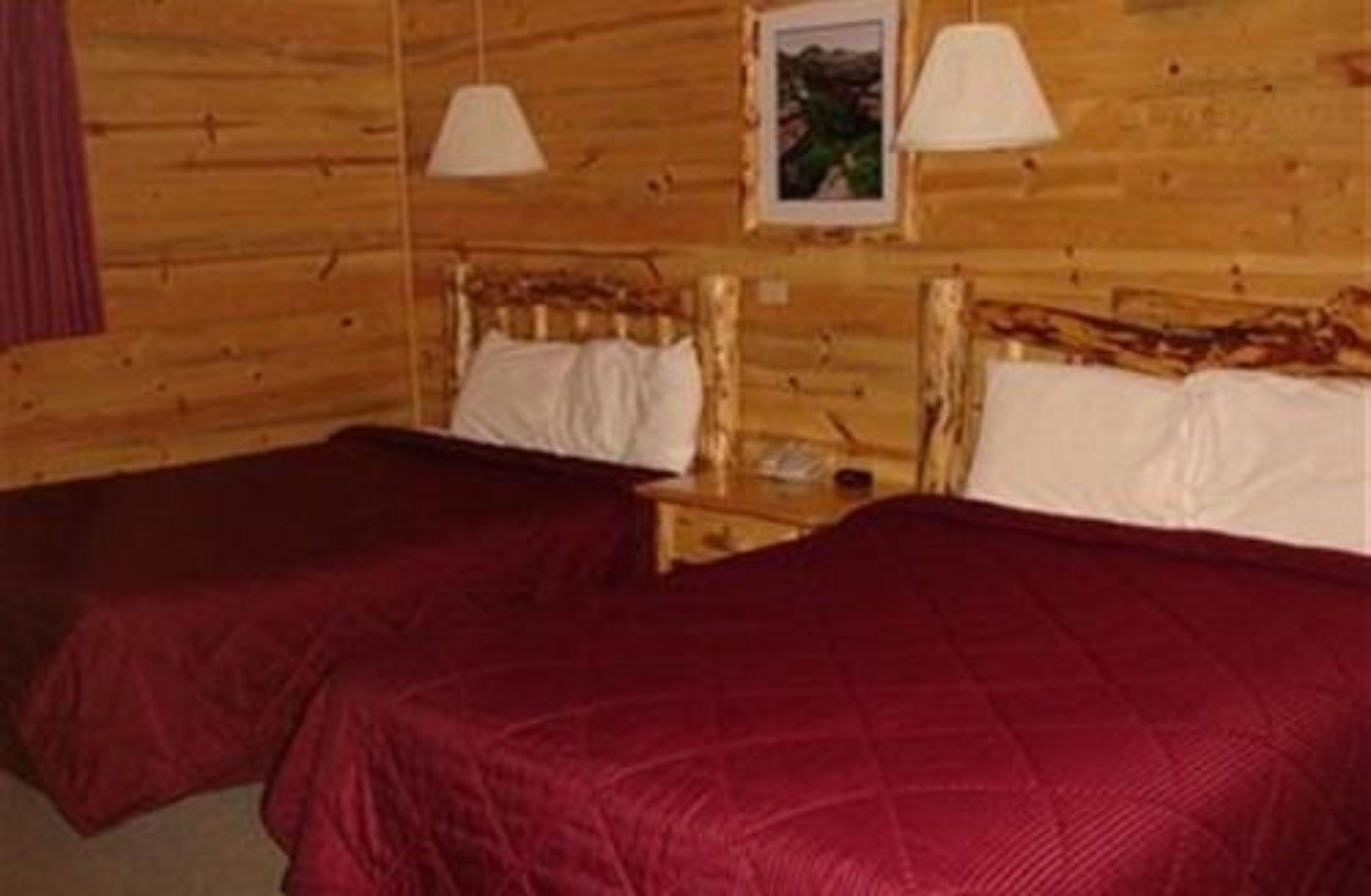 En stuga med träpanel, ett rum med två sängar, var och en med ett rött överkast och vita kuddar. Det finns två sängbord med lampor och en inramad tavla på väggen.