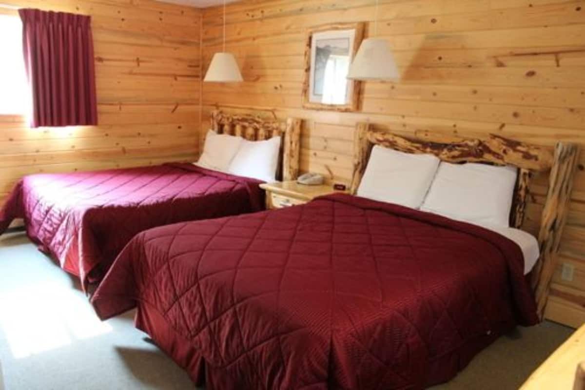 Quarto de cabana de madeira com duas camas, cada uma com colcha vermelha, uma mesa de cabeceira com abajur e uma janela com cortina vermelha.