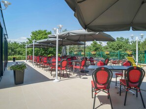 Een terras buiten met tafels en stoelen, afgedekt met parasols, omgeven door groen.