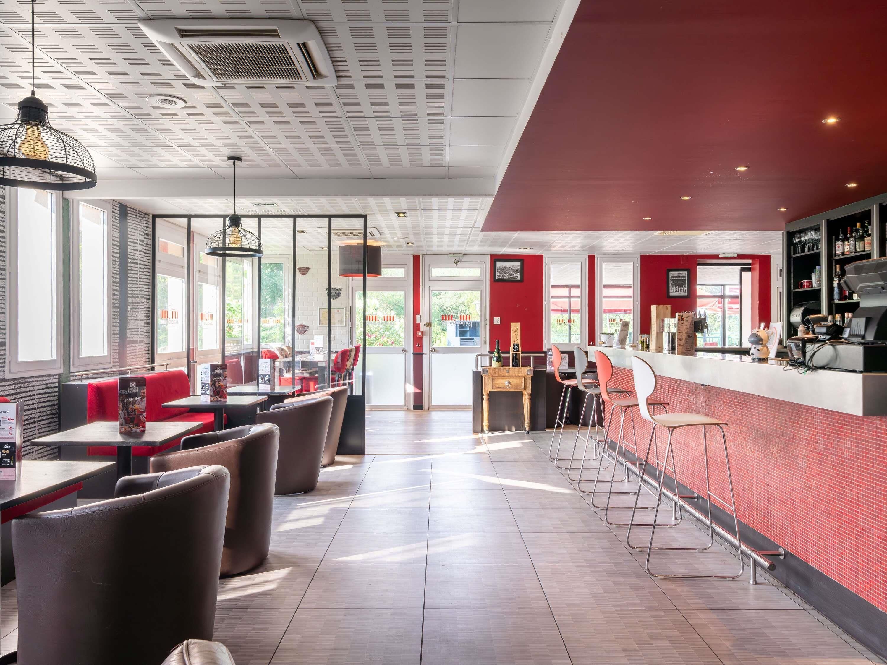 A restaurant interior with high ceilings, large windows, and a red brick accent wall.