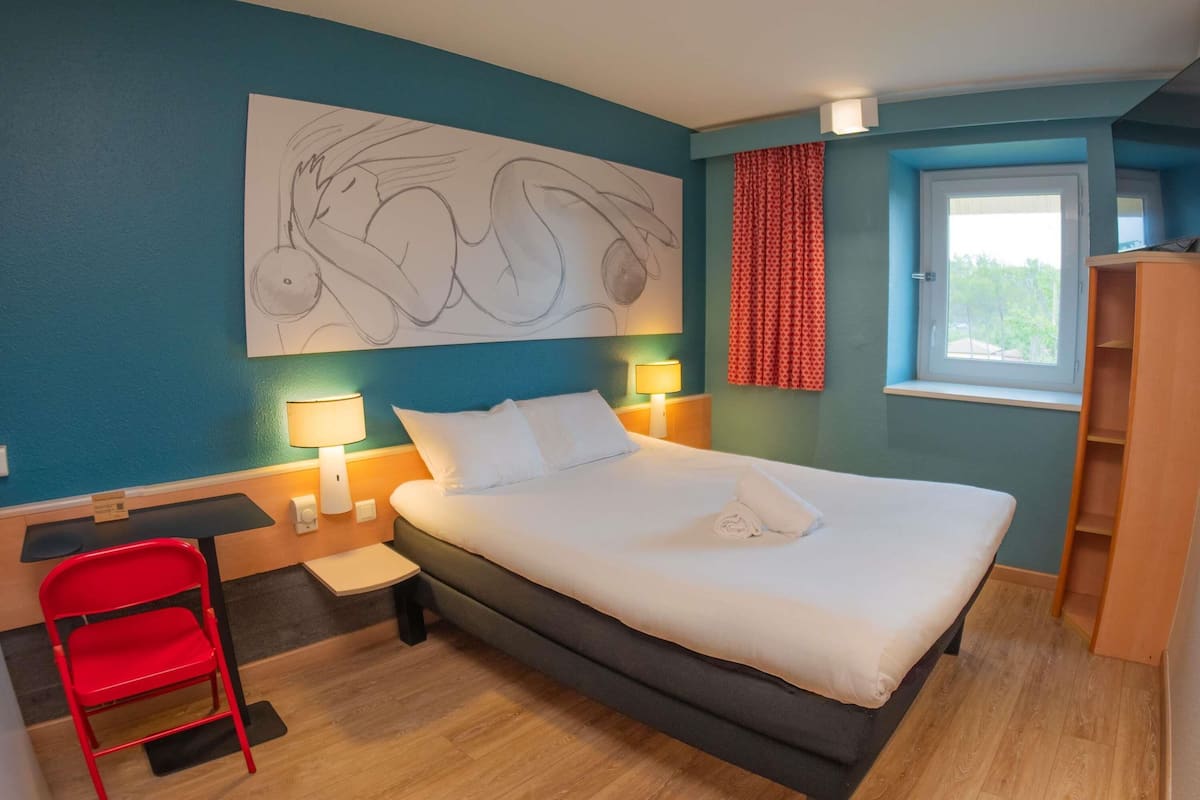 Hotelkamer met een groot bed, een bureau, een stoel, een raam met gordijnen en een kunstwerk aan de muur.