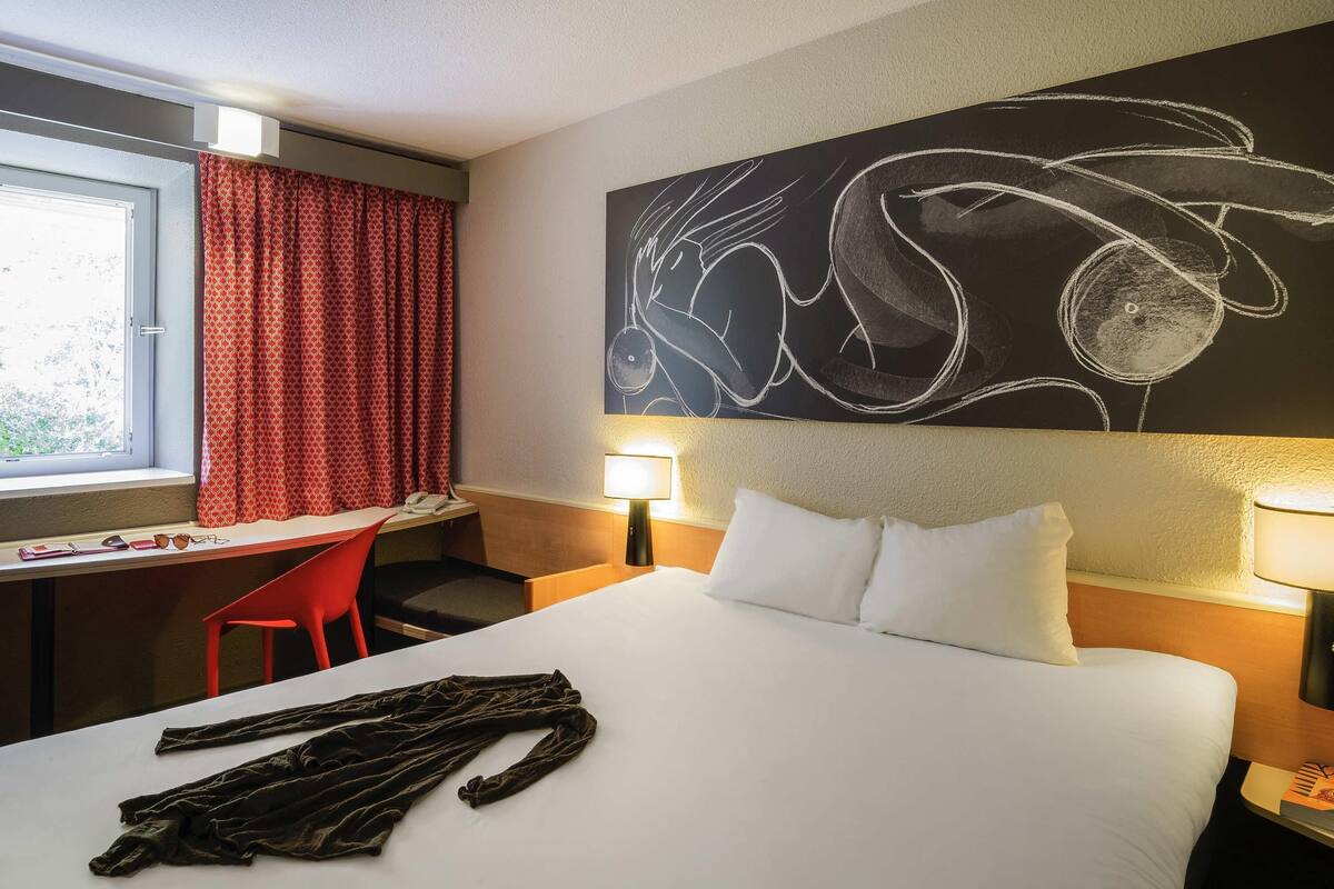 A hotel room with a bed, a desk, a chair, and a large abstract painting on the wall.