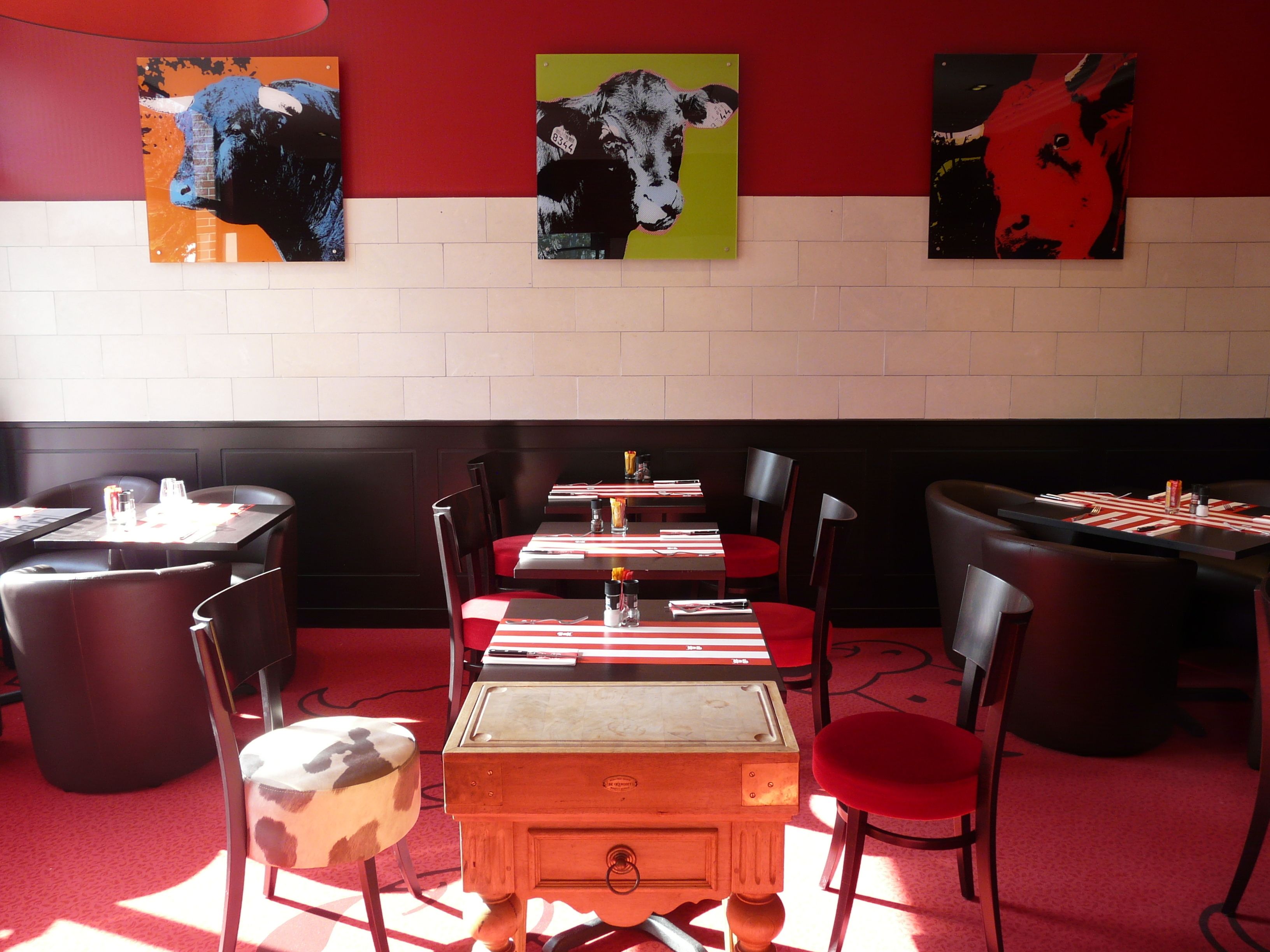 Interno di un ristorante con pareti rosse e bianche, sedie nere e rosse, e tavoli apparecchiati con tovaglioli e posate rosse.
