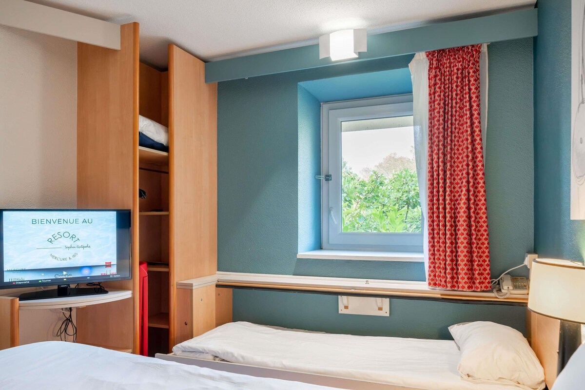 A hotel room with a bed, a desk with a computer, a window with curtains, and a view of greenery outside.