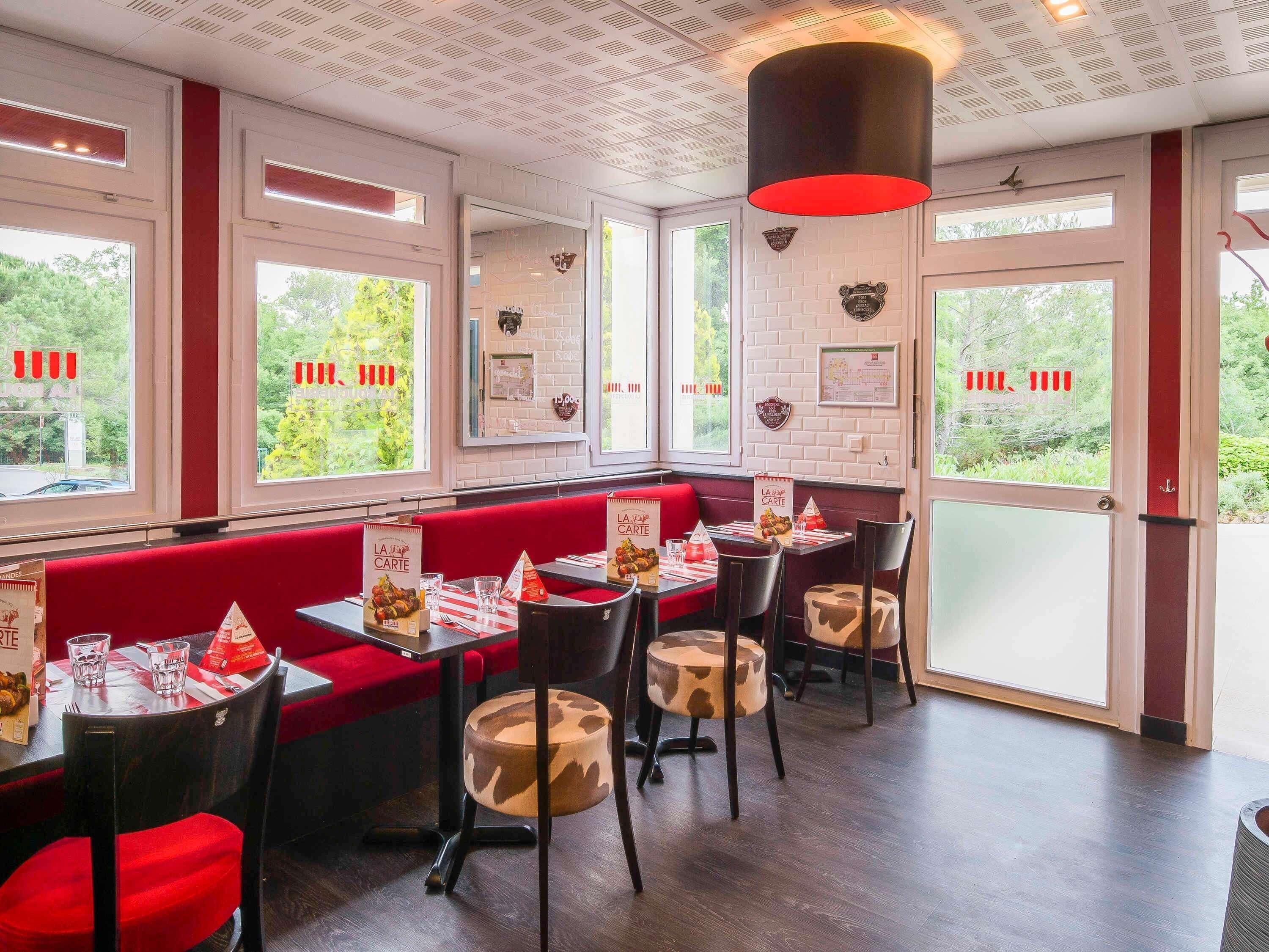 L’intérieur d’un restaurant présente des banquettes rouges, des murs en briques blanches et de grandes fenêtres donnant sur un paysage verdoyant.