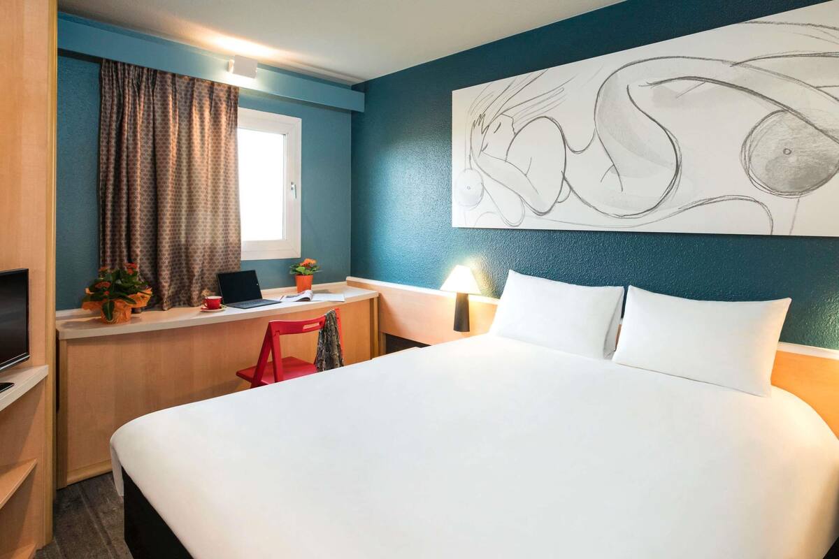 Een hotelkamer met een bed, bureau, televisie en een groot abstract schilderij aan de muur.