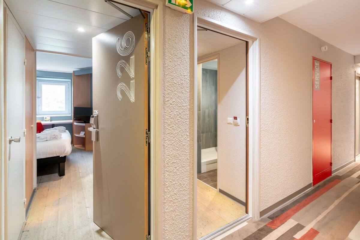 Een hotelgang met een kamer aan de linkerkant, een servicedeur en een badkamer zichtbaar door de deuropening.