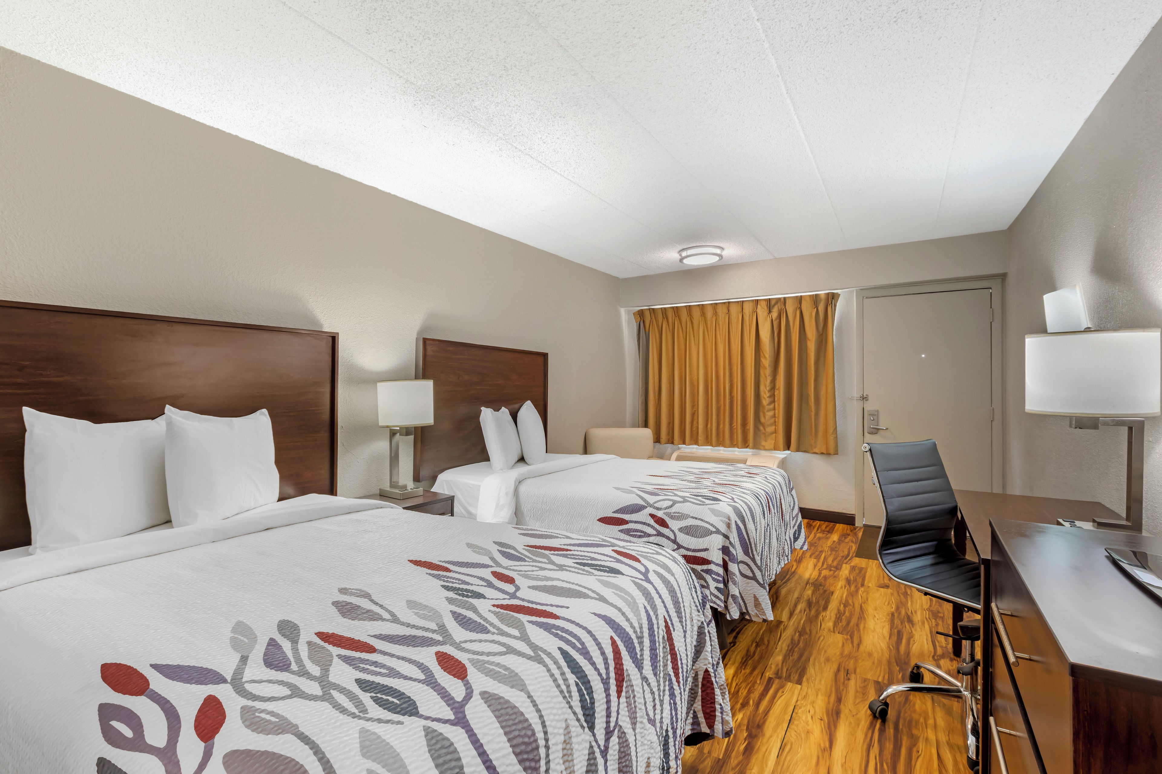 A hotel room with two beds, a desk, a chair, and a bathroom door.