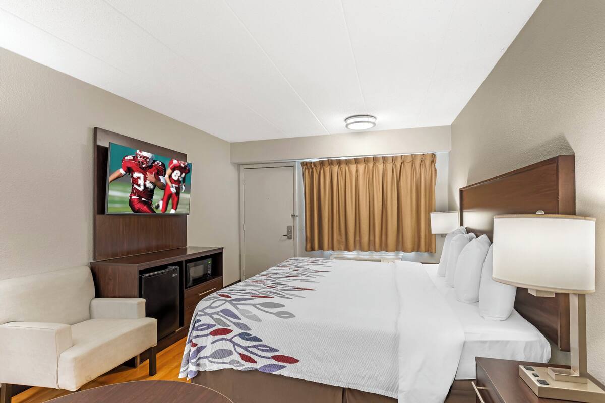 A hotel room with a bed, a TV mounted on the wall, a desk, a chair, and a window with curtains.