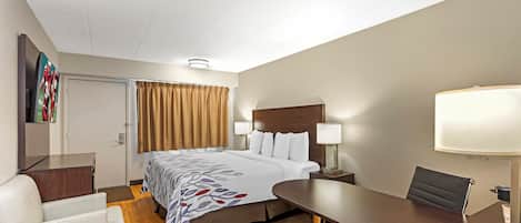 Quarto de hotel com cama, mesas de cabeceira, escrivaninha, cadeira, luminária e televisão na parede.