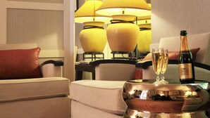 Une chambre d’hôtel avec une table à manger dressée, agrémentée d’une bouteille de champagne et de deux verres, d’un coussin rouge à motifs et d’un fauteuil beige.