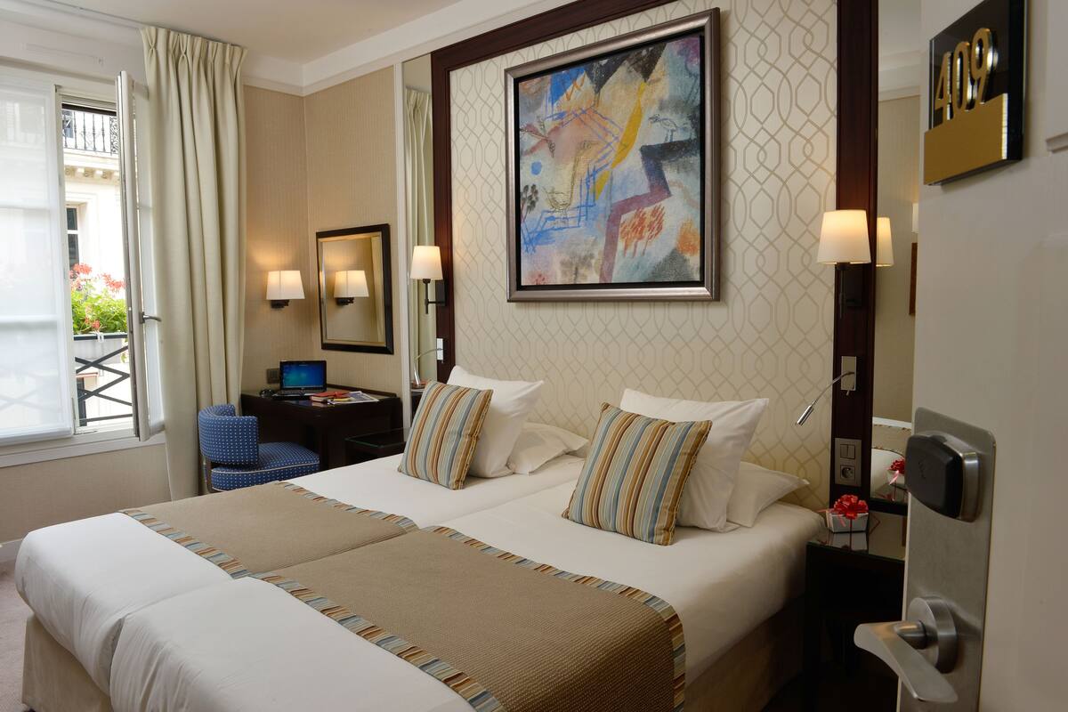 A hotel room with two beds, a desk, a chair, a painting, and a door with a number plate.