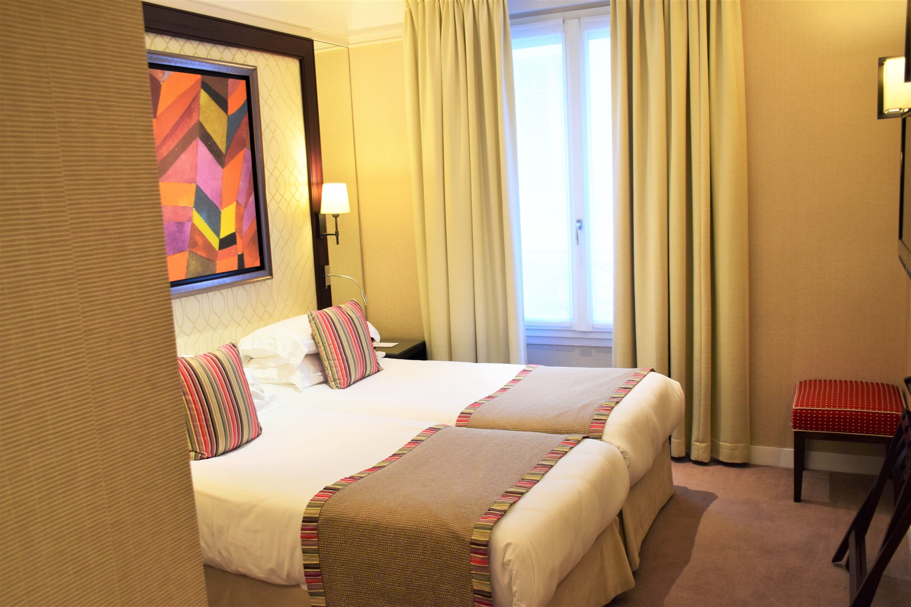 A hotel room with two beds, a small table, a chair, a painting on the wall, and a window with curtains.