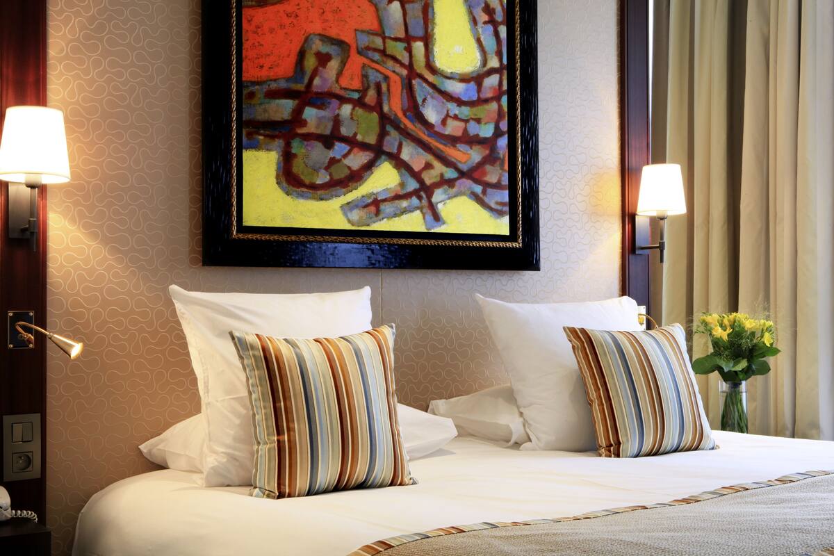 A hotel room with a large bed, two pillows, a framed abstract painting, a vase with flowers, and two wall-mounted lamps.
