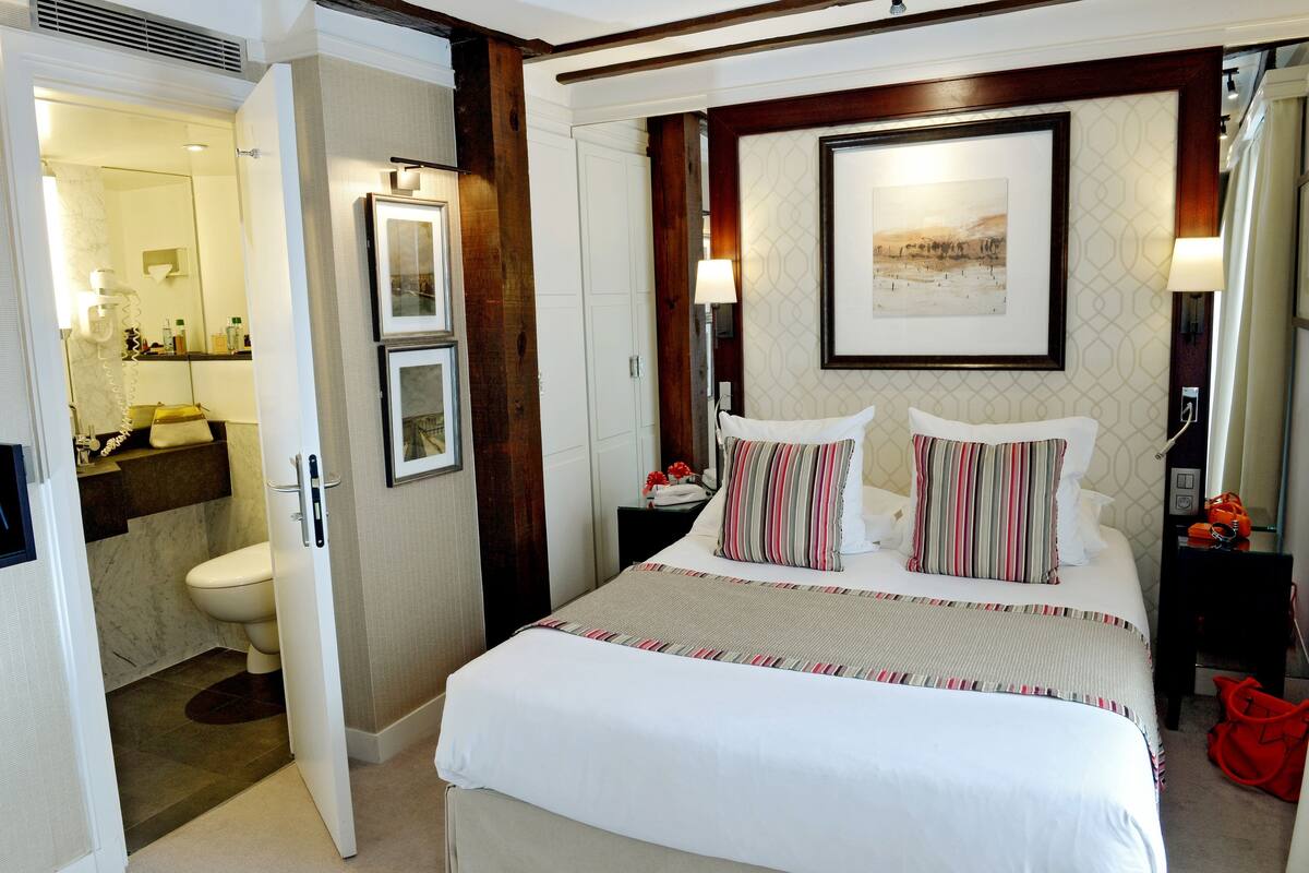 A hotel room with a large bed, a bedside table, a nightstand, a mirror, a framed picture on the wall, and a bathroom visible through an open door.