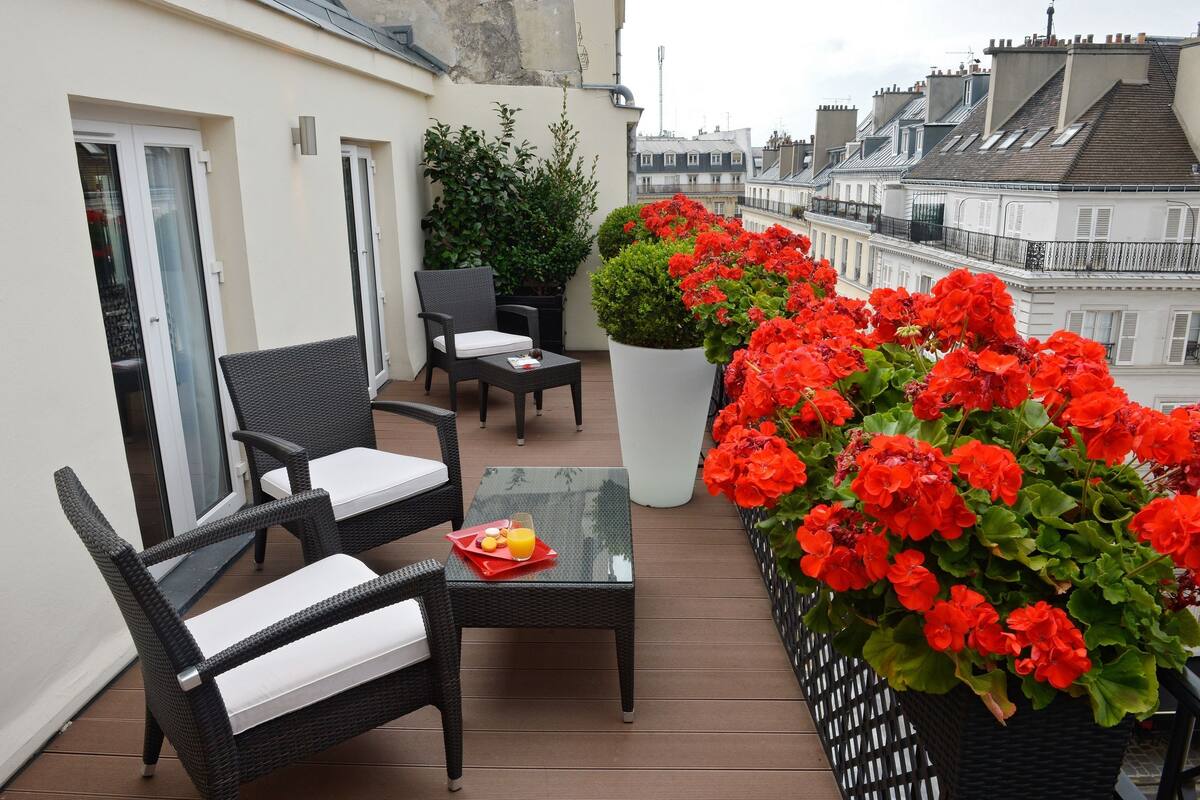 A balcony with wicker furniture, a glass table, and a potted plant with red flowers.