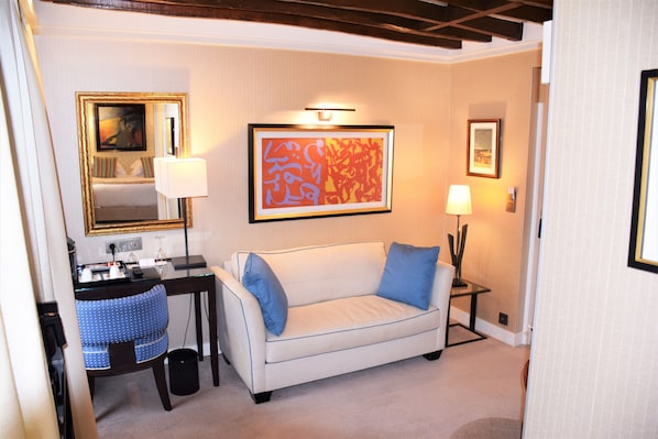 A hotel room with a white sofa, a desk with a chair, a small table with a lamp, and framed artwork on the walls.