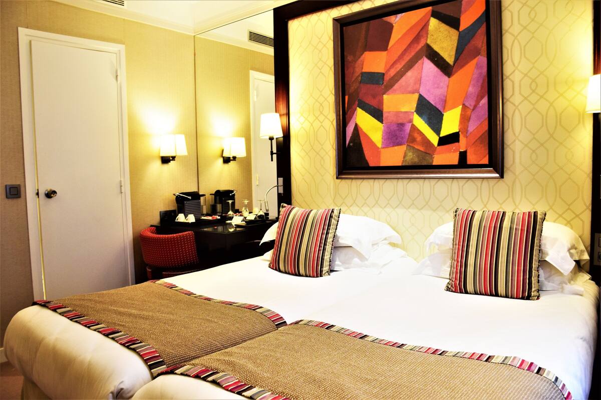 A hotel room with a large bed, two bedside tables, a desk with a chair, and a painting on the wall.