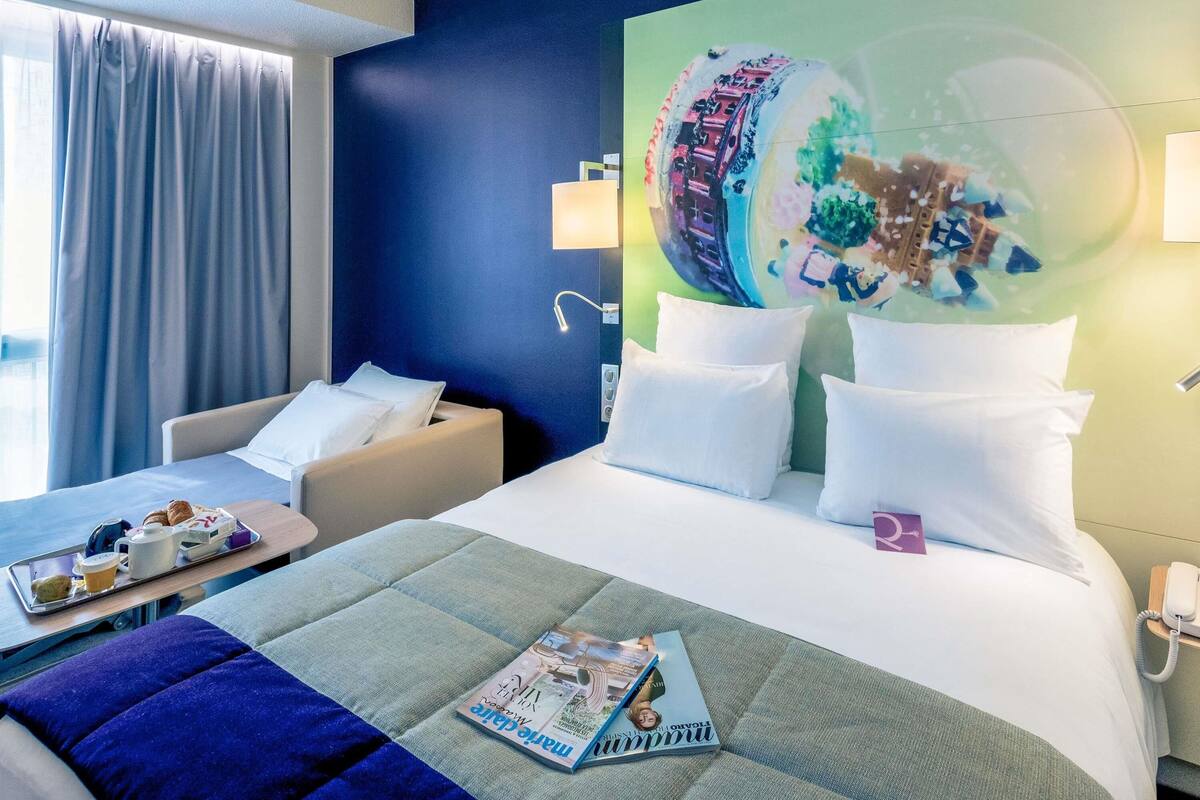 Une chambre d’hôtel avec un lit, un canapé, un plateau contenant des articles pour le petit-déjeuner et un magazine posé sur le lit.
