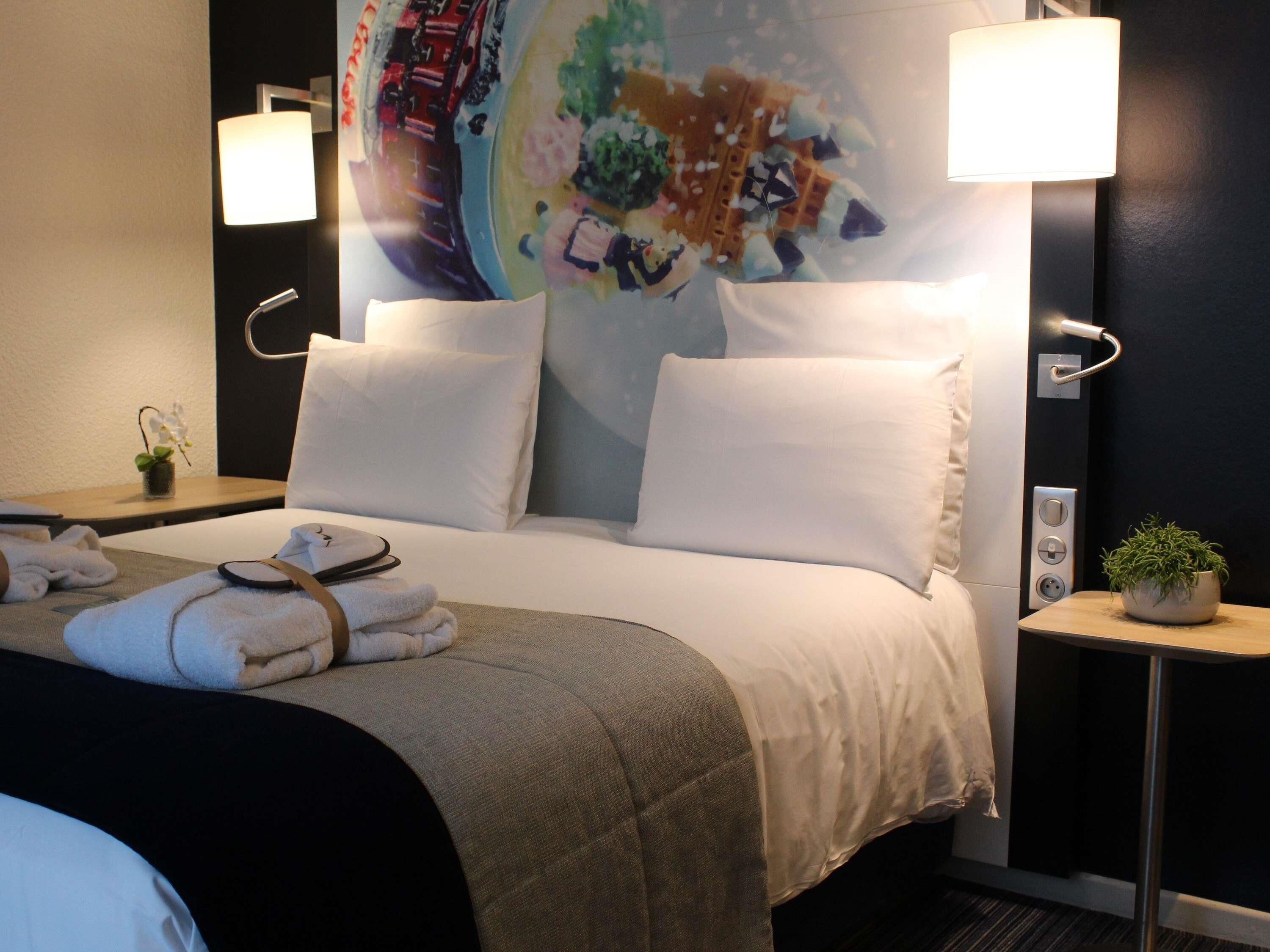 A hotel room with a bed, two bedside lamps, a small table with a plant, and a wall with a colorful mural.