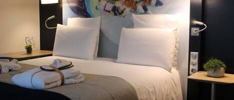 A hotel room with a bed, two bedside lamps, a small table with a plant, and a wall with a colorful mural.