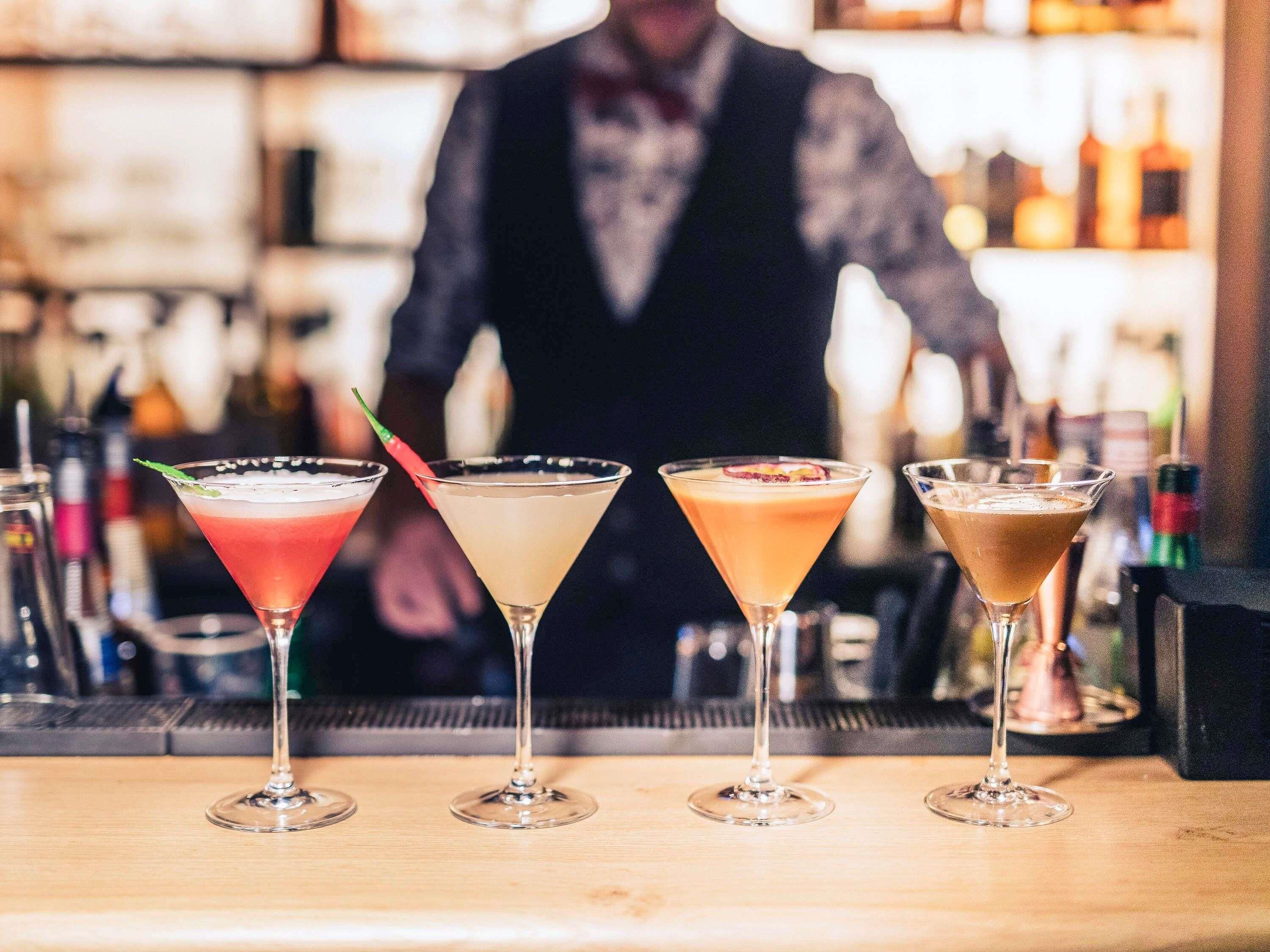Un bar con cuatro vasos de martini de diferentes colores, un cantinero al fondo y una variedad de botellas en estantes.