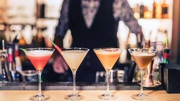 Un bar con cuatro vasos de martini de diferentes colores, un cantinero al fondo y una variedad de botellas en estantes.