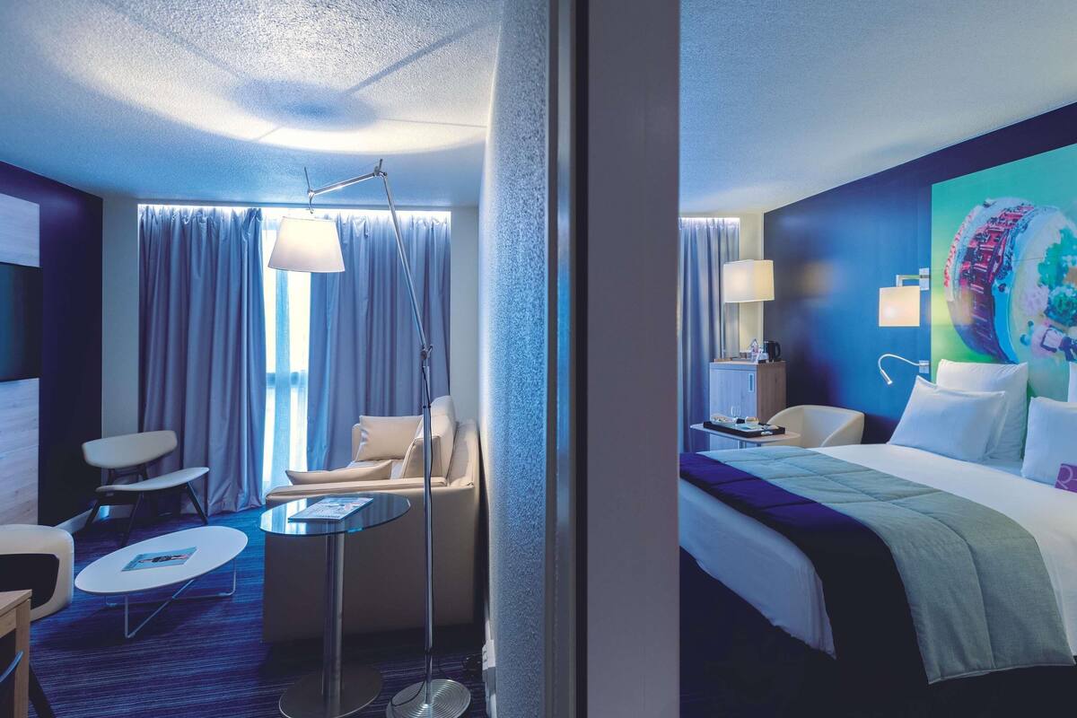 Une chambre d’hôtel moderne équipée d’un lit, d’un bureau, d’une chaise, d’une petite table et d’une lampe sur pied.