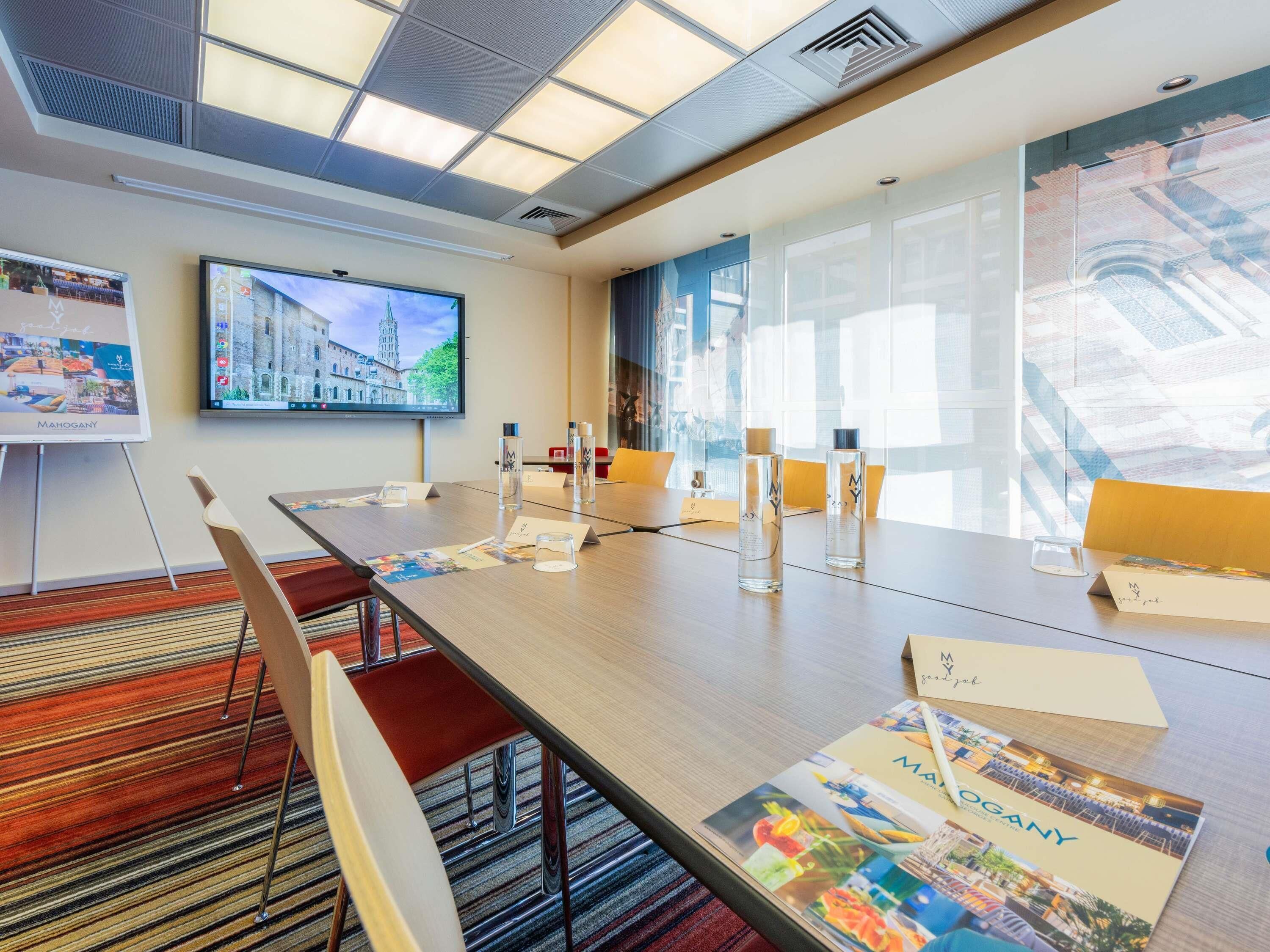 A conference room with a long table, chairs, a projector screen, and a large window with a view of buildings.