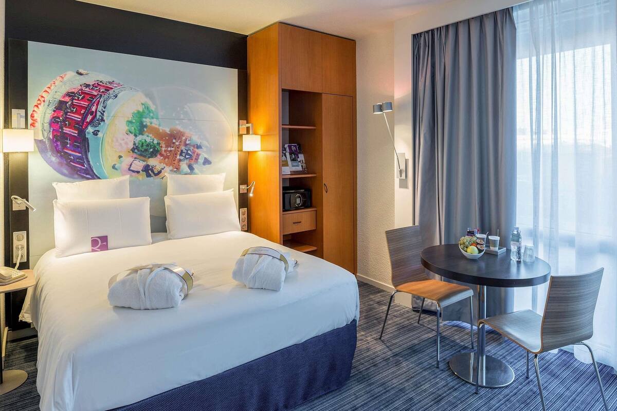 Ein Hotelzimmer mit einem großen Bett, einem Schreibtisch, einem Stuhl, einem kleinen Tisch mit Obstschale und einem Bild an der Wand.