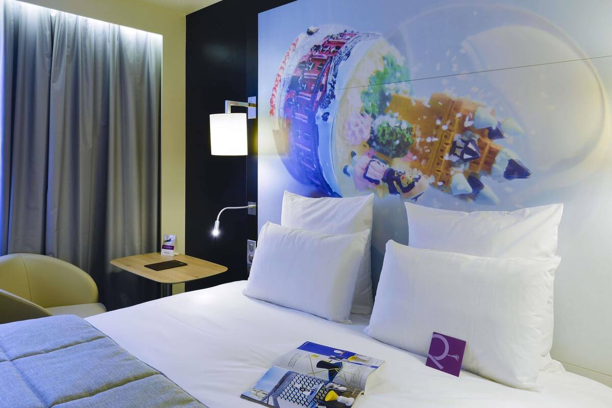 Ein Hotelzimmer mit Bett, Nachttisch, Lampe, Stuhl und einem großen Wandbild.