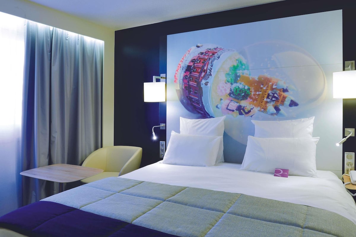 Ein Hotelzimmer mit einem großen Bett, einem Nachttisch, einem Sessel und einer Wand mit einem farbenfrohen Wandbild.