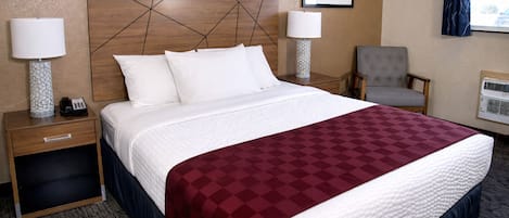 A hotel room with a large bed, two bedside tables with lamps, a chair, a framed picture on the wall, and an air conditioning unit.