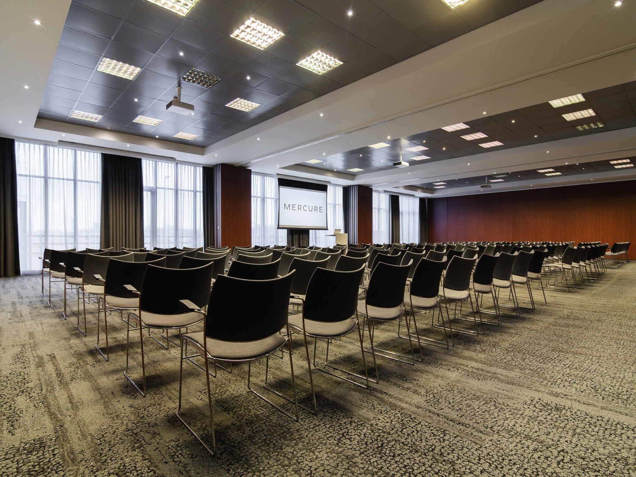 A conference room with rows of chairs, a large screen displaying 'MERCURE', and a projector mounted on the ceiling.