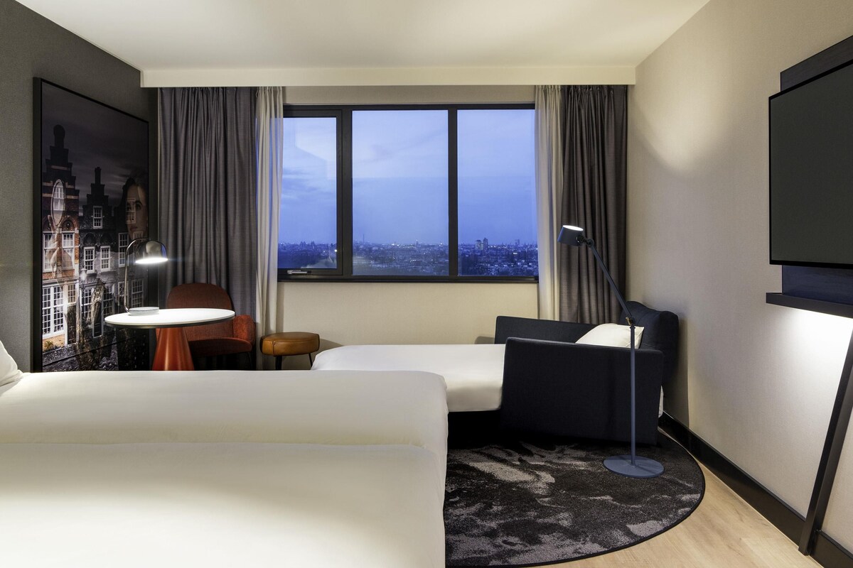 A hotel room with a bed, a desk, a chair, a TV, and a window with a city view.