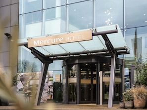 A entrada do Hotel Mercure, com fachada de vidro, uma placa sobre a entrada e uma porta giratória.