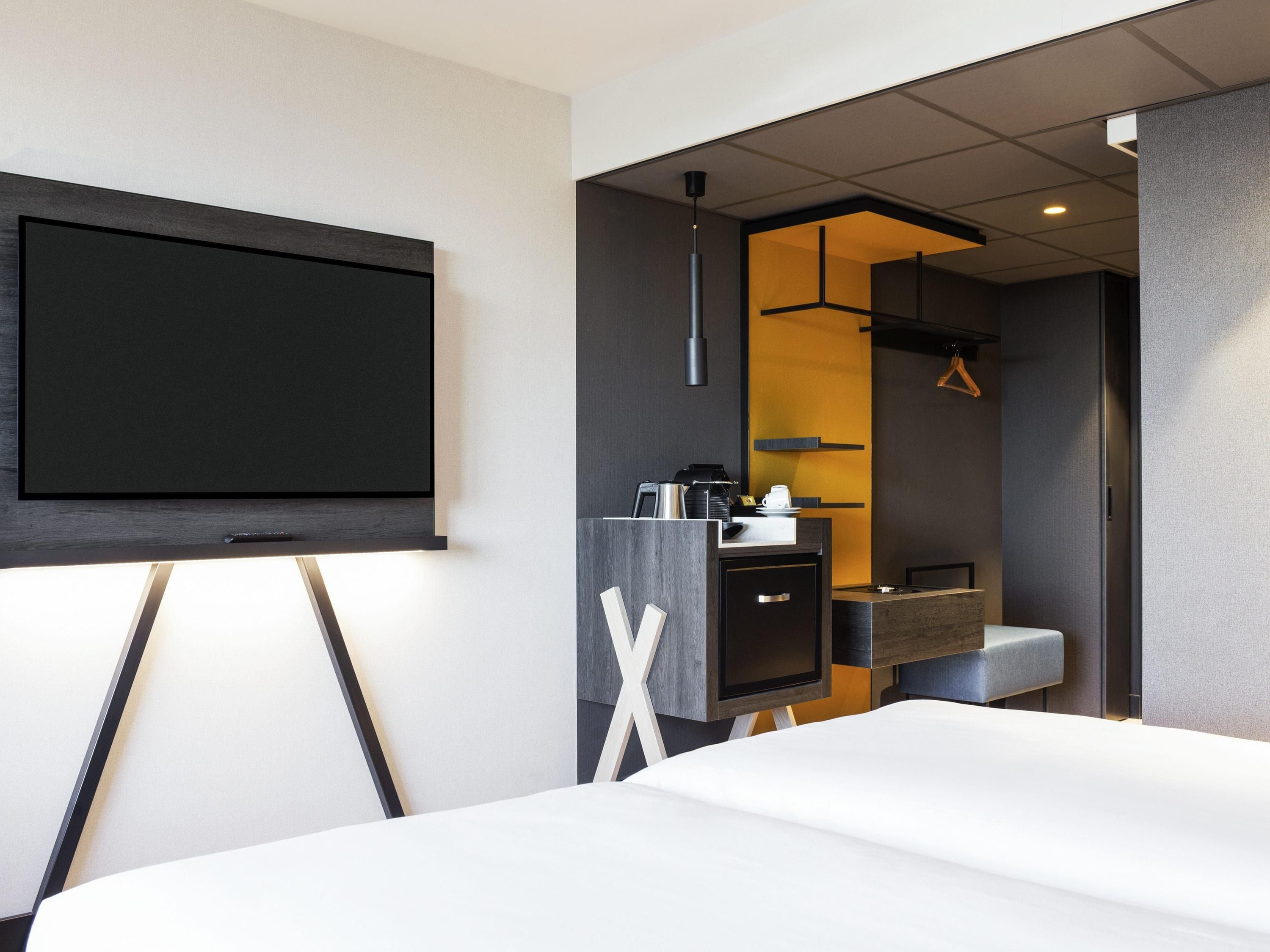 A modern hotel room with a large flat-screen TV mounted on the wall, a bed with white bedding, a small desk with a coffee maker, and a mirror above the sink.
