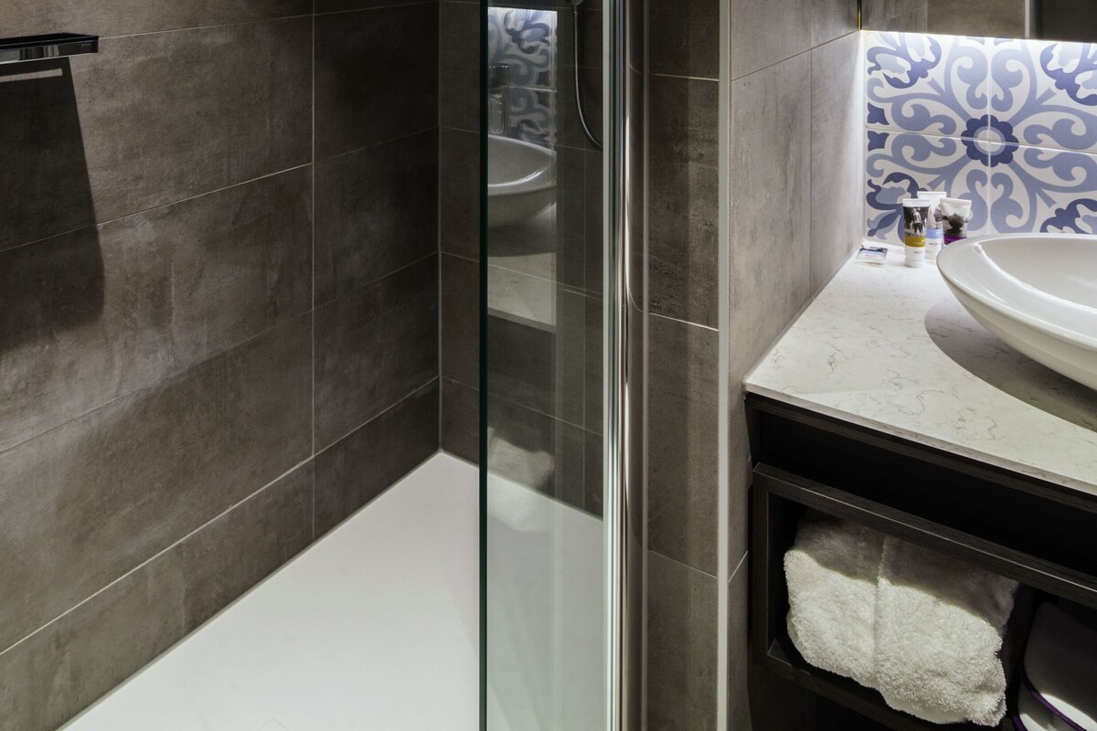 Un baño moderno con ducha de vidrio, paredes cubiertas de azulejos oscuros, un lavamanos blanco y un salpicadero de azulejos con diseño.