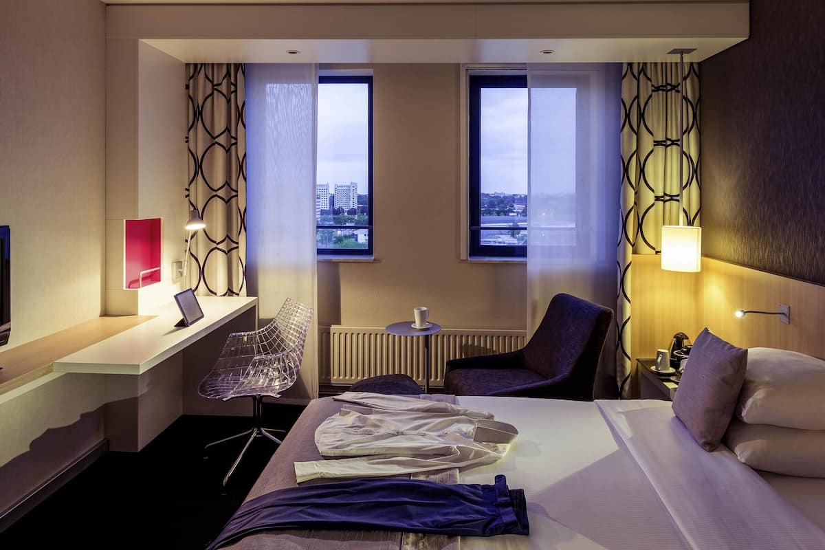 Una habitación de hotel con cama, escritorio, silla, mesa y dos ventanas.