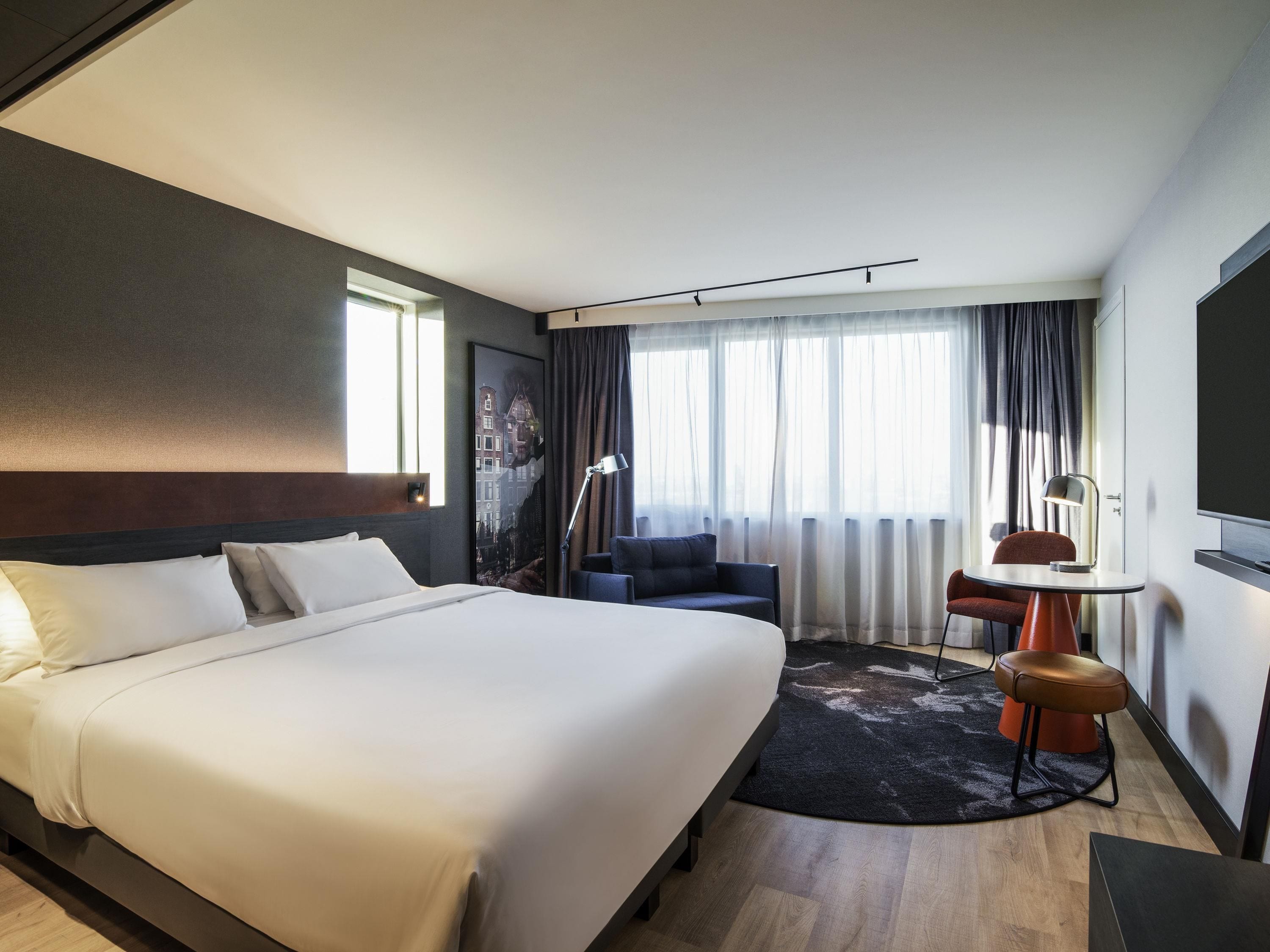A hotel room with a large bed, a desk, a chair, and a view of the city through the window.