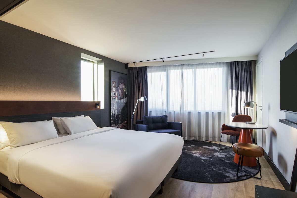 Habitación de hotel con una cama grande, un escritorio, una silla y vista a la ciudad por la ventana.
