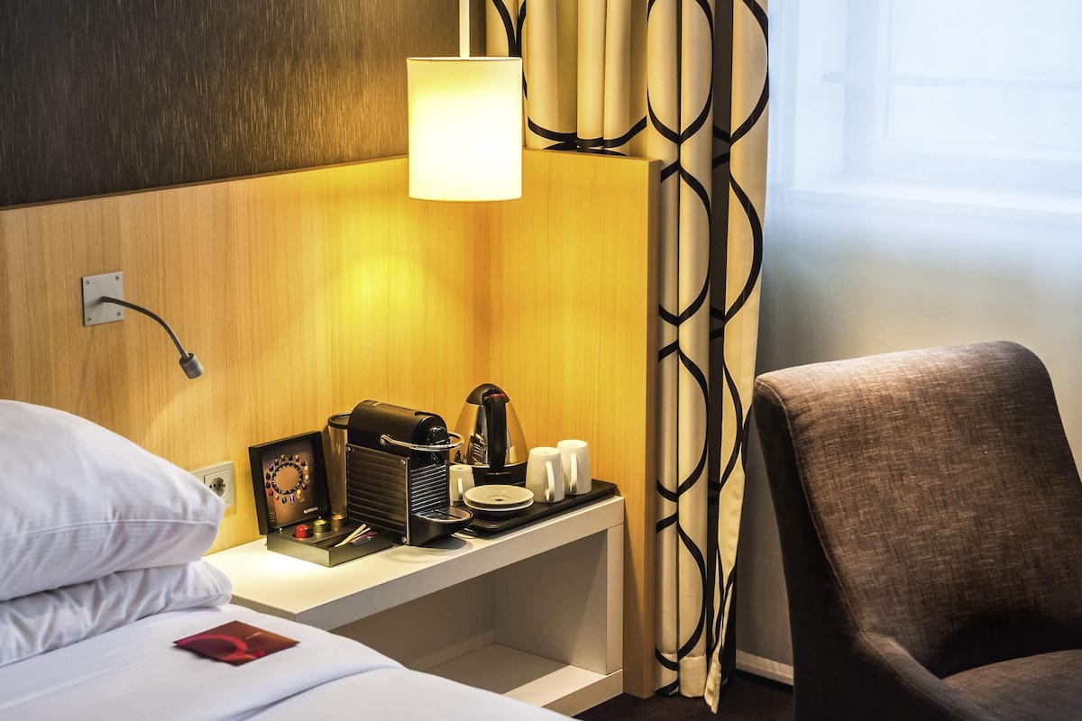 Habitación de hotel con cama, escritorio con lámpara, cafetera y una silla.