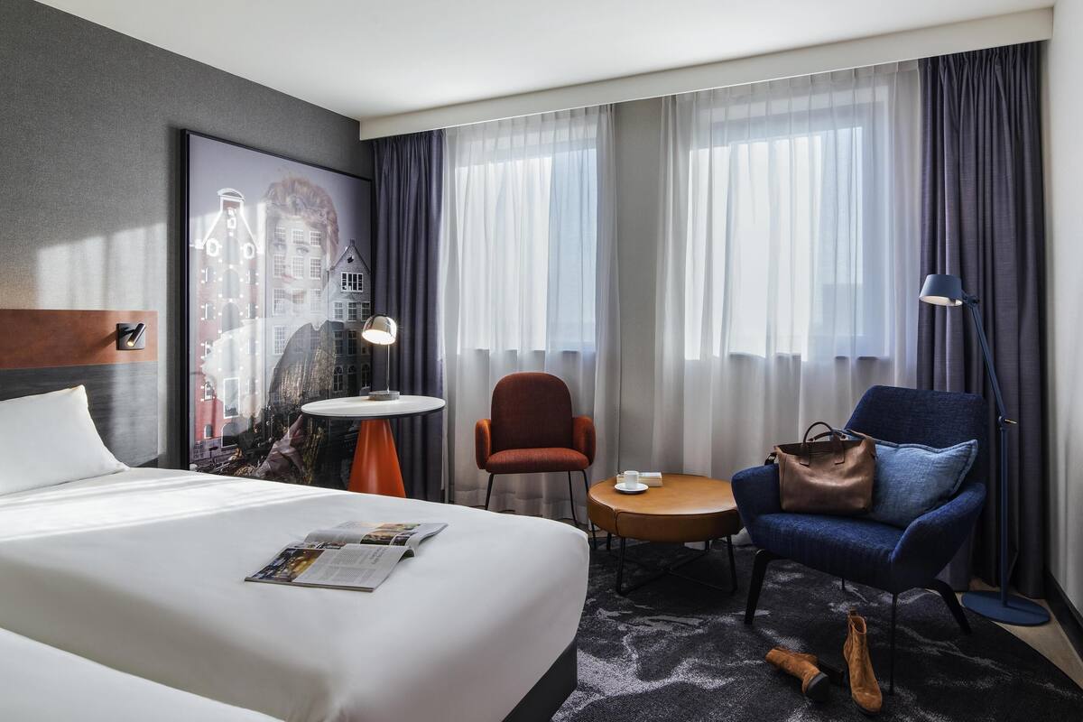 Una habitación de hotel con una cama grande, una mesita redonda pequeña, un sillón azul, una lámpara de pie y un ventanal con cortinas.