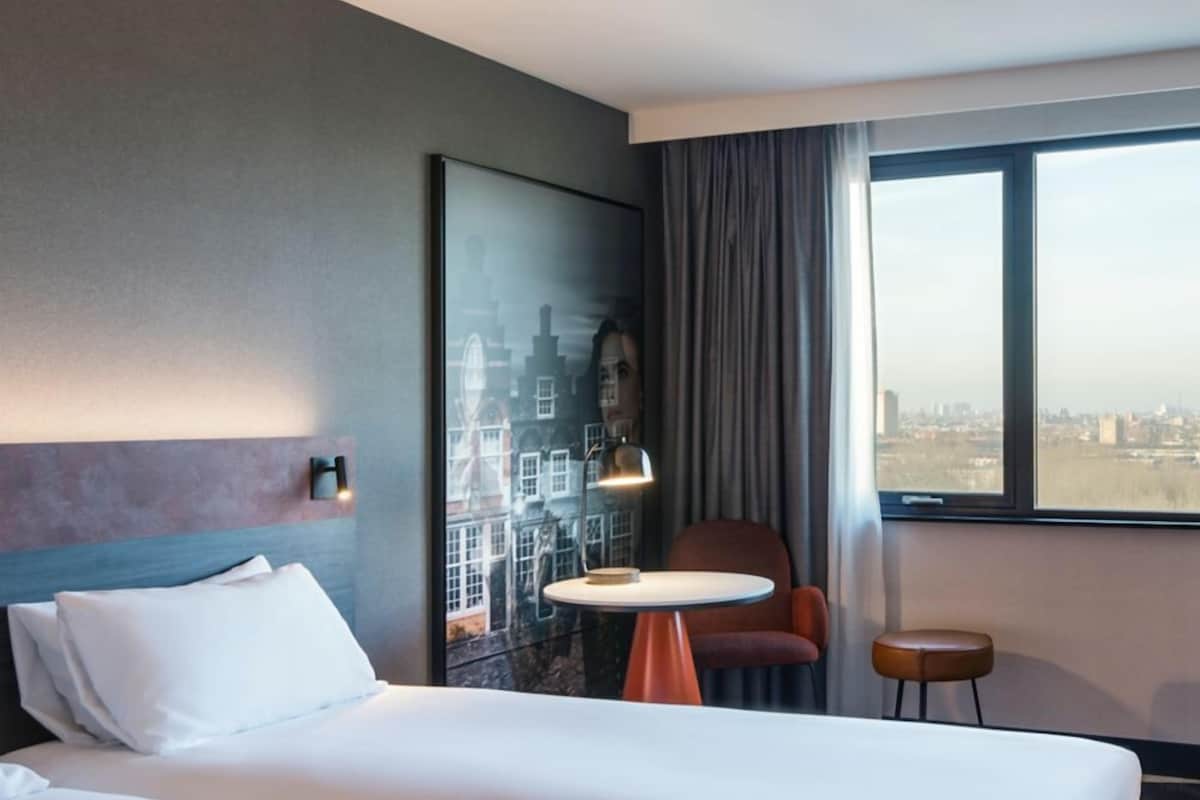 Habitación de hotel con cama, una mesita, una silla y vistas de la ciudad a través de un ventanal grande.