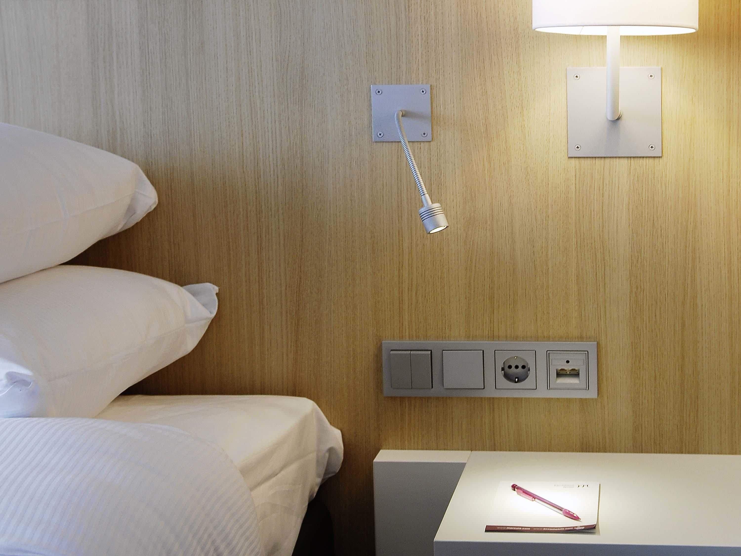A hotel room with a bed, a nightstand, a lamp, and a power outlet.