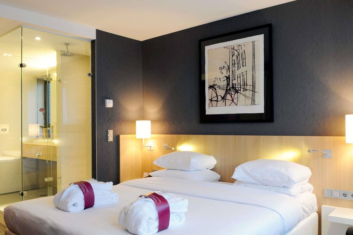 Habitación de hotel con una cama, dos lámparas de pared, una obra de arte enmarco y cabina de ducha con mampara de vidrio.