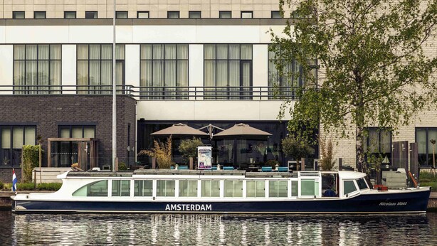 A boat named 'AMSTERDAM' docked at a waterfront location with a building in the background.